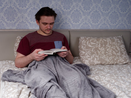 a young guy in a red T-shirt sits on a light-colored bed and reads a thick book, the concept of rest after a working dayの写真素材