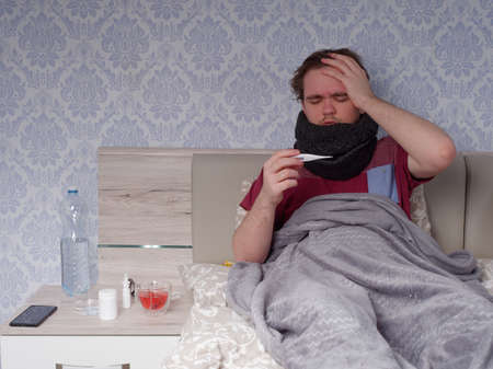 a young guy in a red T-shirt sits on the bed and measures his temperature with an electronic thermometer, the concept of illness at homeの写真素材