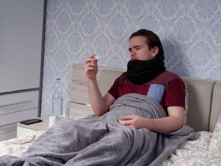 a young guy in a red T-shirt sits on the bed and measures his temperature with an electronic thermometer, the concept of illness at homeの写真素材