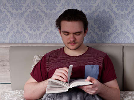 a young guy in a red T-shirt sits on a light-colored bed and reads a thick book, the concept of rest after a working dayの写真素材