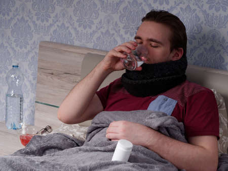 young guy in a red t-shirt sits on the bed and takes cough pills, pain relievers and cold medications, the concept of illness at homeの写真素材