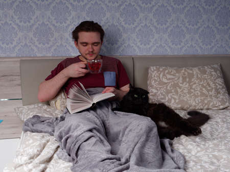 a young guy in a red T-shirt sits on a light-colored bed and reads a thick book, the concept of rest after a working dayの写真素材