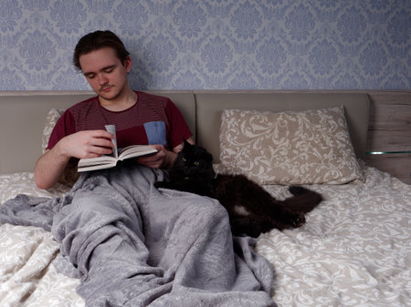 a young guy in a red T-shirt sits on a light-colored bed and reads a thick book, the concept of rest after a working dayの写真素材
