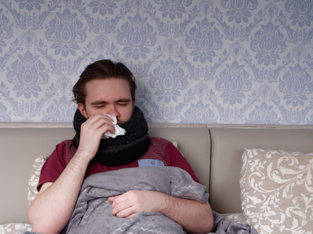 young guy in a red T-shirt sits on the bed and takes care of his sore nose, he has a runny nose, the concept of illness at homeの写真素材