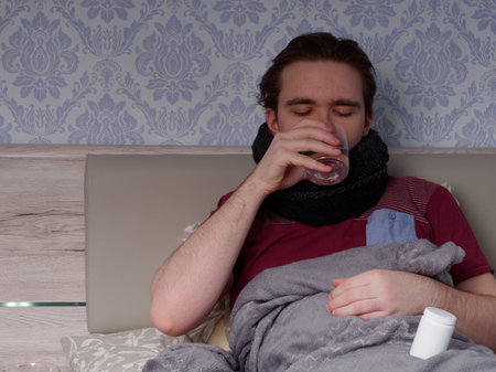young guy in a red t-shirt sits on the bed and takes cough pills, pain relievers and cold medications, the concept of illness at homeの写真素材