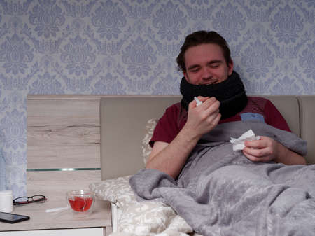 young guy in a red T-shirt sits on the bed and takes care of his sore nose, he has a runny nose, the concept of illness at homeの写真素材