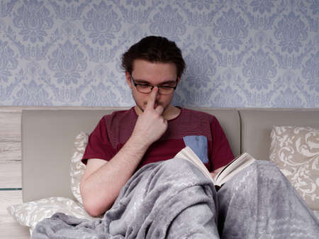 a young guy in a red T-shirt sits on a light-colored bed and reads a thick book, the concept of rest after a working dayの写真素材