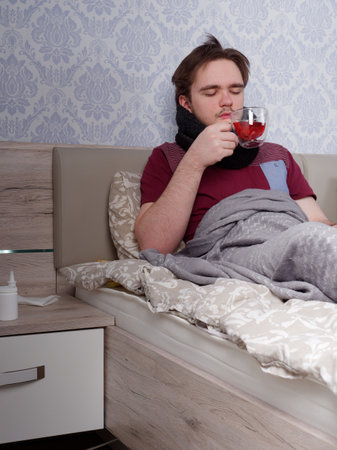 a young guy in a red T-shirt sits on the bed and drinks tea for a cold, the concept of illness at homeの写真素材
