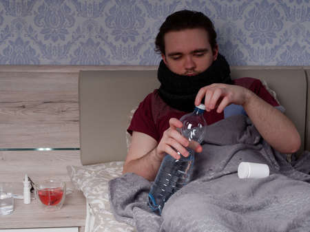 young guy in a red t-shirt sits on the bed and takes cough pills, pain relievers and cold medications, the concept of illness at homeの写真素材