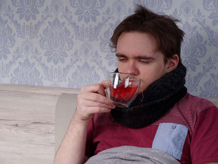 a young guy in a red T-shirt sits on the bed and drinks tea for a cold, the concept of illness at homeの写真素材