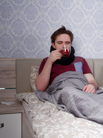 a young guy in a red T-shirt sits on the bed and drinks tea for a cold, the concept of illness at homeの写真素材