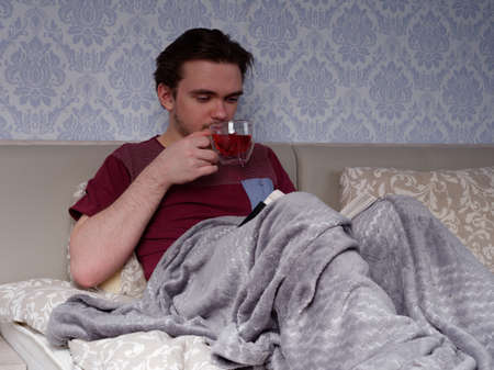 a young guy in a red T-shirt sits on a light-colored bed and reads a thick book, the concept of rest after a working dayの写真素材