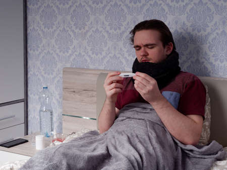 a young guy in a red T-shirt sits on the bed and measures his temperature with an electronic thermometer, the concept of illness at homeの写真素材