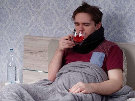 a young guy in a red T-shirt sits on the bed and drinks tea for a cold, the concept of illness at homeの写真素材