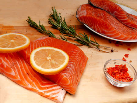 Close-up of two slices of fresh raw salmon fillets on a white plate and two on a wooden cutting board along with rosemary and spices. Healthy eating conceptの写真素材