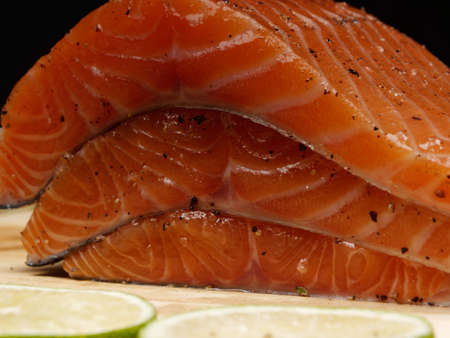 A close-up of three slices of fresh raw red salmon fillets which lie on top of each other, and in front of them a green lemon, lie on a wooden cutting board. Healthy food conceptの写真素材