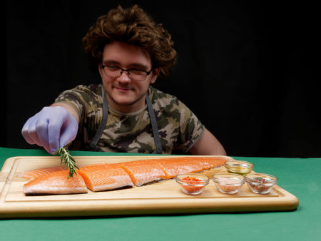 A young chef puts rosemary on a fresh raw salmon fillet which lies next to spices on a wooden...