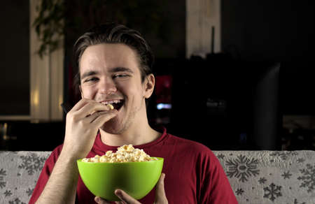 A young guy in a red T-shirt sits on the couch on which a white blanket, and eats popcorn and smiles while watching TV. Weekend and relaxation conceptの写真素材