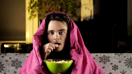 A young guy in a red T-shirt is sitting on the couch under a pink blanket, eating popcorn and watching TV intently. Weekend and relaxation conceptの写真素材