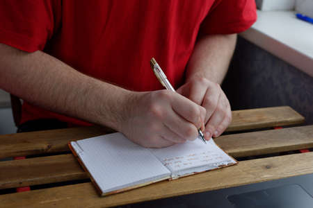 A young guy in a red T-shirt sits at the table, holds a pen in his hands and writes in a notebookの写真素材