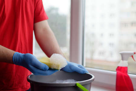A young guy in a red T-shirt holds a yellow sponge on which there is foam over a bucket of water. Window cleaner conceptの写真素材