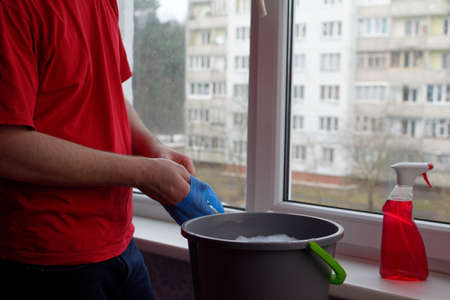 A young guy in a red T-shirt puts on a blue glove and stands next to a bucket of water and window cleaner. Window cleaner conceptの写真素材
