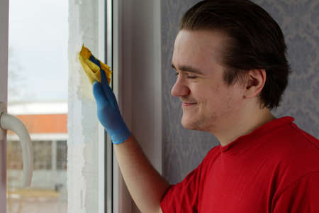 A young guy in a red T-shirt and blue gloves holds a yellow rag in his hands and wipes the window. Window cleaner conceptの写真素材