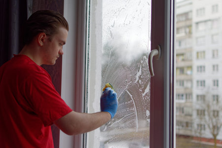 A young guy in a red T-shirt holds a yellow sponge in his hands and washes the window. Window...