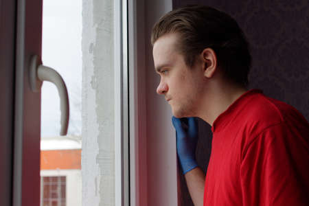 A young guy in a red T-shirt stands leaning against the wall and looks out the window at the street.の写真素材