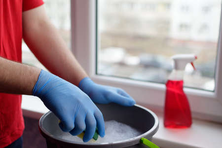 A young guy in a red T-shirt holds a sponge in his hands and is going to wet it in water for cleaning windows. Window cleaner conceptの写真素材