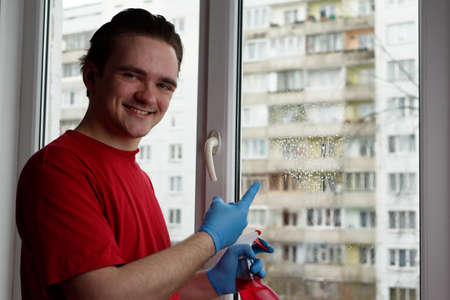 A young guy in a red T-shirt holds a bottle with a red window cleaner in his hands, and points with his finger to the window where the liquid is. Window cleaner conceptの写真素材