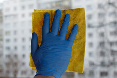 A male hand in blue gloves holds a yellow rag on the glass. Window cleaner concept