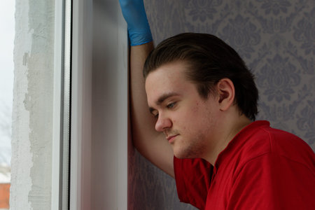 A young guy in a red T-shirt stands leaning against the wall and looks out the window at the street.