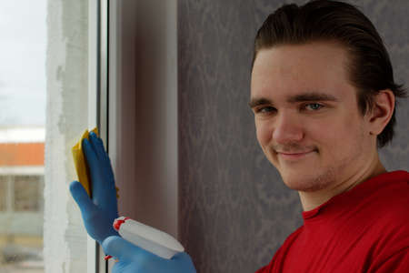 A young guy in a red T-shirt and blue gloves holds a yellow rag and a bottle of red liquid in his hands and wipes the window. Window cleaner conceptの写真素材