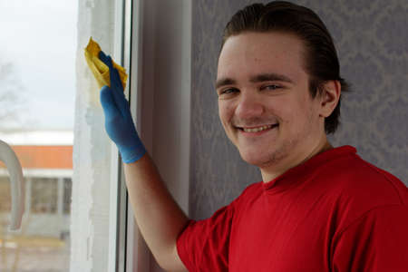 A young guy in a red T-shirt and blue gloves holds a yellow rag in his hands and wipes the window. Window cleaner conceptの写真素材