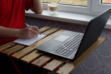 A young guy in a red T-shirt sits at the table, holds a pen in his hands and writes in a notebook, in front of him is a laptop and a cup of coffee on the windowsillの写真素材