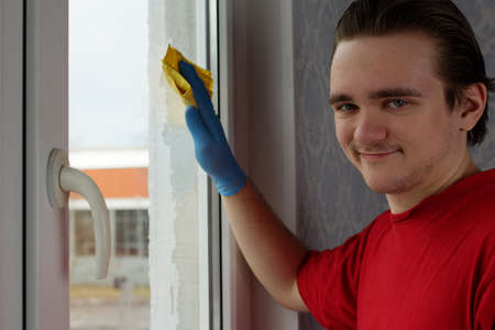A young guy in a red T-shirt and blue gloves holds a yellow rag in his hands and wipes the window. Window cleaner conceptの写真素材