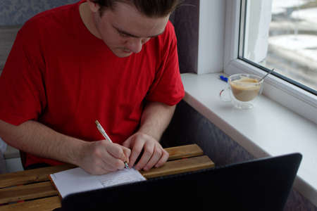 A young guy in a red T-shirt sits at the table, holds a pen in his hands and writes in a notebook, in front of him is a laptop and a cup of coffee on the windowsillの写真素材