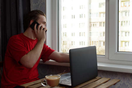 A young guy in a red T-shirt sits at the table and talks on the phone, next to there is a laptop and a cup of coffeeの写真素材