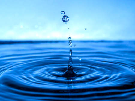 A blue drop drips into the water and creates splashes of various shapes, waves are created through the water, the concept of a liquid splash, a substance painted in blue colorの写真素材