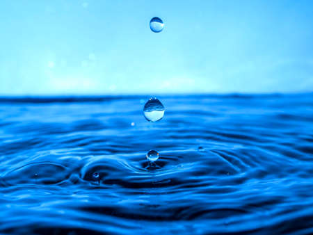 A blue drop drips into the water and creates splashes of various shapes, waves are created through the water, the concept of a liquid splash, a substance painted in blue colorの写真素材