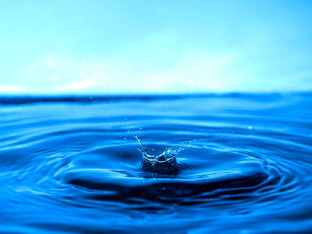 A blue drop drips into the water and creates splashes of various shapes, waves are created through the water, the concept of a liquid splash, a substance painted in blue colorの写真素材
