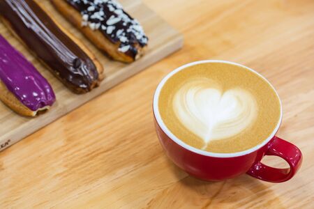 Red cup of hot art cappuccino with a heart on a foam. Assorted eclairs on the table.の写真素材