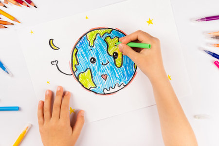 Child girl draws planet earth with multicolored felt-tip pens on a white sheet.