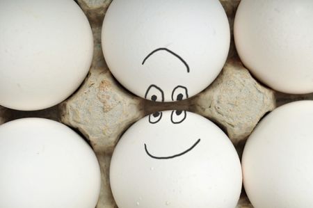 Closeup of six white new eggs in a cardboard egg carton. Two of the eggs in the middle have drawn on faces and are looking at each other upside down.の写真素材