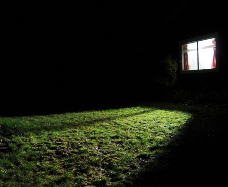 A dark house at night with a window with a light on in the room spilling out light onto the grass lawn yardの写真素材