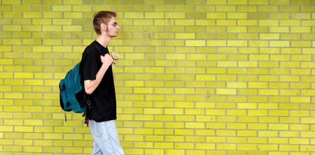 A tall white Caucasian young adult teenage male walks in front of a brick wall. Lots of room for your copyspace text. He has a backpack and glassesの写真素材