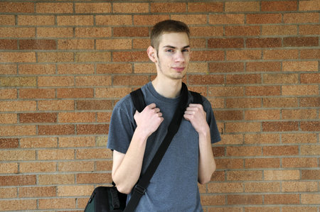 Young caucasian male looks at the camera with his backpack on against a wall.