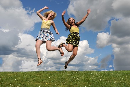 Two girls jumping over a grass hill with their arms up in the air below the blue sky with white clouds.の写真素材