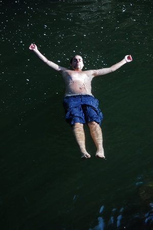 A young man swims and floats peacefully on his back in calm dark green lake water